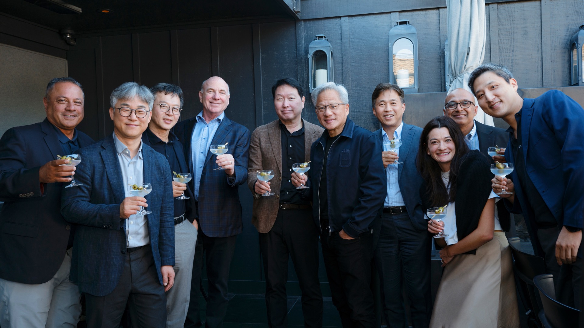A group of ten people pose for a photo at an outdoor social event, holding cocktail glasses with olives; they wear business attire against a dark wood-paneled backdrop with lantern-style wall lights.